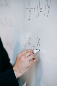 An educator uses a marker to solve equations on a whiteboard, demonstrating math skills.