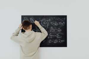 Teenager in hoodie writing math formulas on a blackboard indoors, showcasing problem-solving skills.